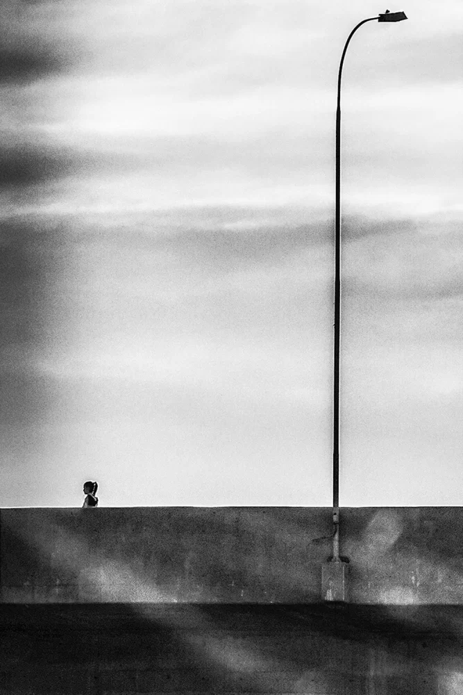 Foto em P&B de Ricardo Laf mostrando uma passarela urbana no Centro de BH, com um poste de iluminação alto e uma pequena silhueta de uma pessoa caminhando solitária.