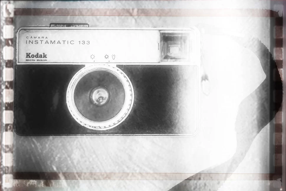 Fotografia em alto contraste de uma câmera Kodak Instamatic 133, apresentada com texturas que remetem a um negativo de filme antigo, em tons de cinza e branco.