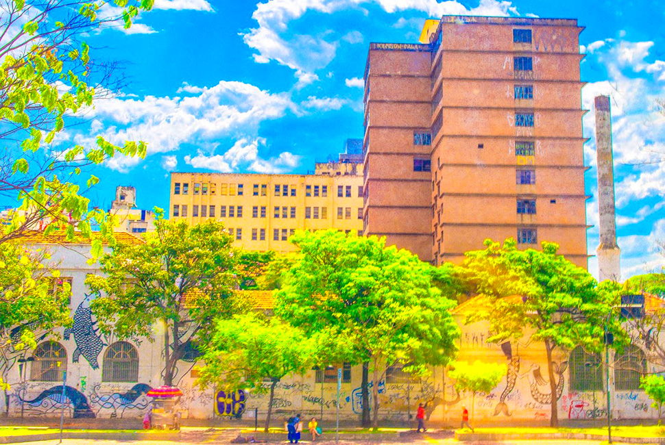 Fotografia de Ricardo Laf mostrando um grande edifício de tijolos e concreto em Belo Horizonte, com árvores em primeiro plano e um céu azul límpido sob luz solar forte.