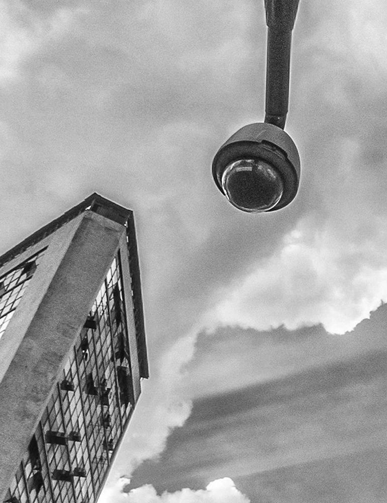 Fotografia em preto e branco de Ricardo Laf mostrando uma câmera de segurança em destaque contra um céu nublado e a silhueta de um edifício no Centro de Belo Horizonte.