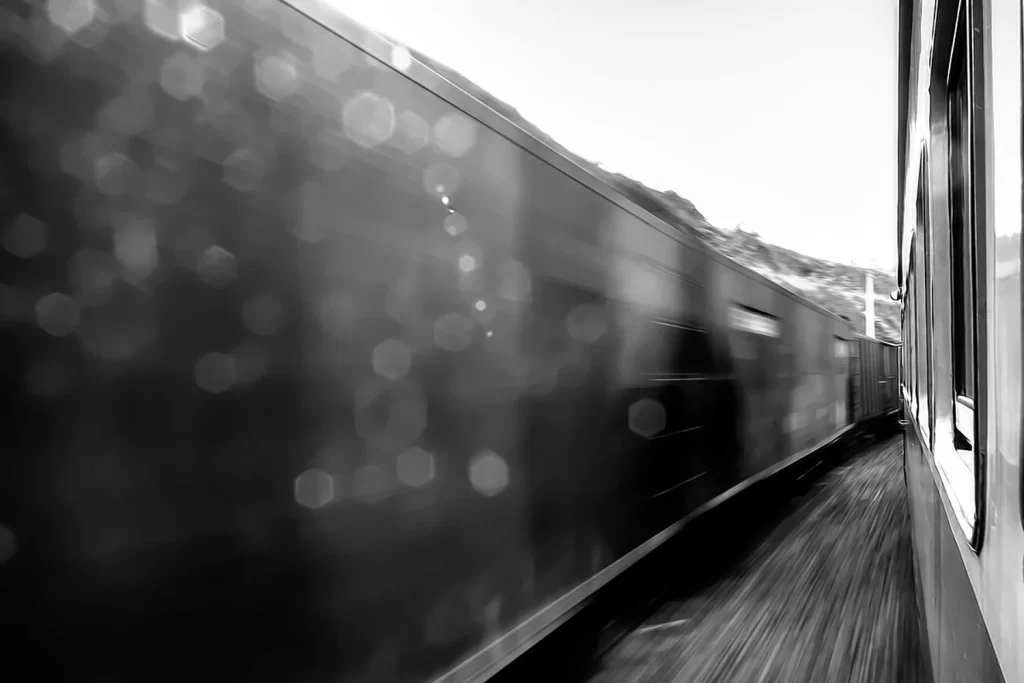 Fotografia em preto e branco de Ricardo Laf através da janela de um trem em movimento, com gotas de chuva no vidro e o cenário externo em desfoque.