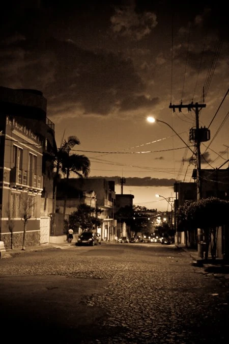 Fotografia de Ricardo Laf capturando a descida de uma rua arborizada na Região Leste de BH, com luzes e sombras projetadas que criam uma atmosfera introspectiva e urbana.