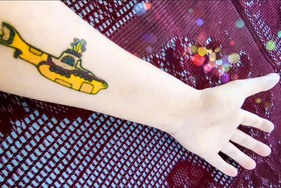Fotografia em close do braço esquerdo de Ricardo Laf, exibindo uma tatuagem colorida inspirada na arte da capa do álbum Yellow Submarine, dos Beatles. A tatuagem mostra o submarino amarelo com detalhes vibrantes sobre a pele.