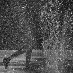 Fotografia autoral em preto e branco de Ricardo Laf mostrando a silhueta de uma pessoa caminhando por trás dos jatos de água da fonte na Praça da Estação, em Belo Horizonte.
