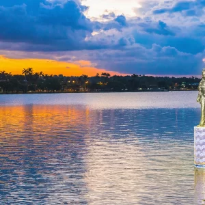 Fotografia de Ricardo Laf mostrando a estátua de Iemanjá na Lagoa da Pampulha, Belo Horizonte, durante o entardecer com luzes da cidade ao fundo.