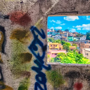 Fotografia autoral de Ricardo Laf mostrando uma vista urbana de Belo Horizonte através de uma abertura retangular em uma parede de concreto com grafite, revelando casas coloridas ao fundo.