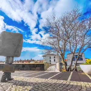 Fotografia de Ricardo Laf capturando a escultura do Monumento ao Burocrata Desconhecido em Reykjavik, Islândia, com um bloco de pedra no lugar da cabeça e tronco, sob um céu azul com nuvens espessas.