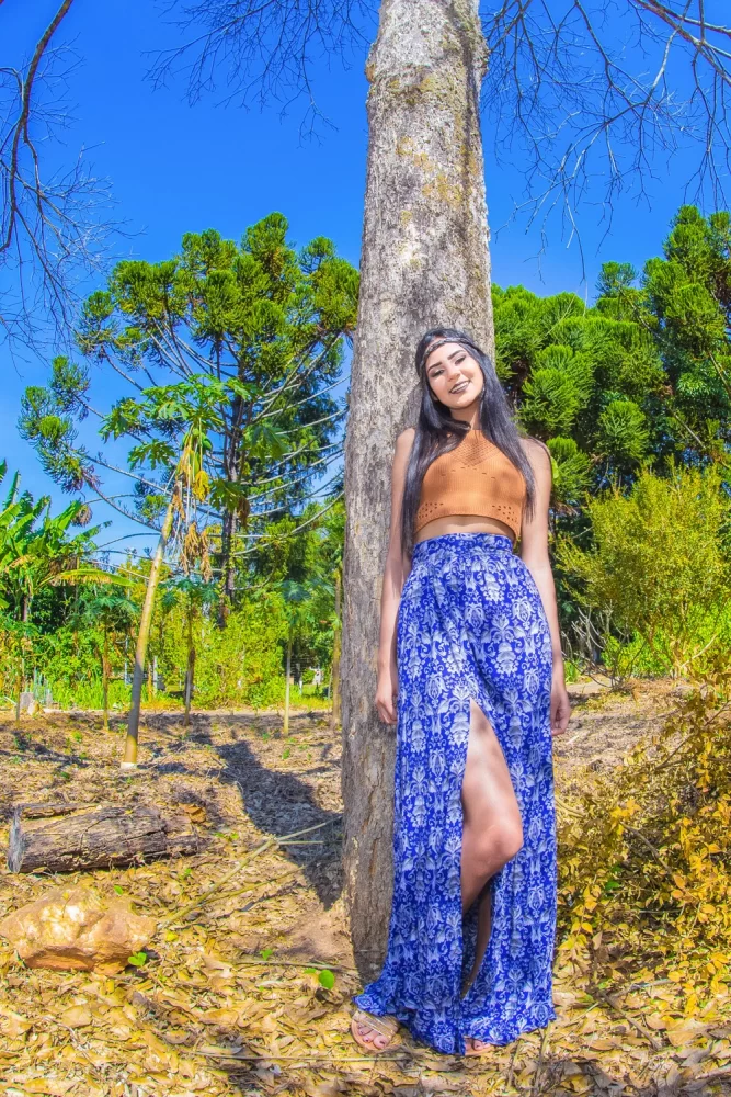 Fotografia autoral de Ricardo Laf em plano inteiro vertical; a modelo Daphne posa encostada em um tronco de árvore, vestindo uma saia azul estampada e top marrom, sob um céu azul sem nuvens.