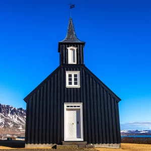 Fotografia de Ricardo Laf da Igreja Negra de Búðir (Búðakirkja) na península de Snæfellsnes, Islândia, destacando a arquitetura em madeira escura contra montanhas nevadas.