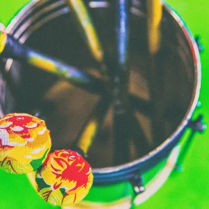 Fotografia de Ricardo Laf em close-up mostrando baquetas coloridas repousando sobre um tambor, com um fundo verde vibrante, capturada na Região Leste de Belo Horizonte.