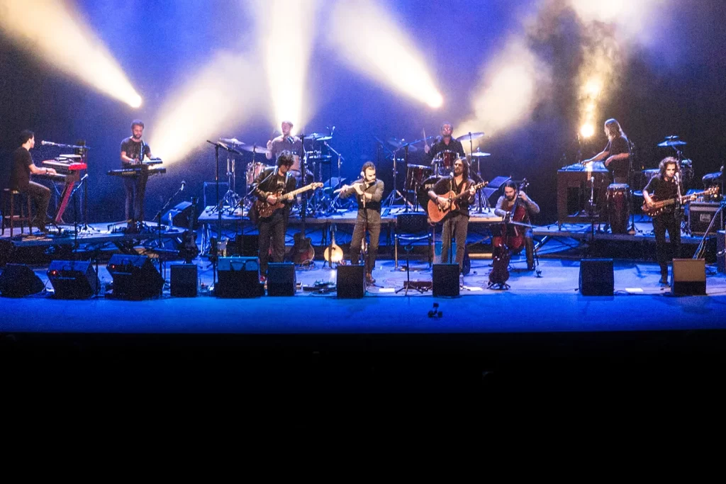 Fotografia em plano médio da Orquestra Mineira de Rock no Sesc Palladium. Músicos no palco sob iluminação lateral dramática em tons de azul e branco.