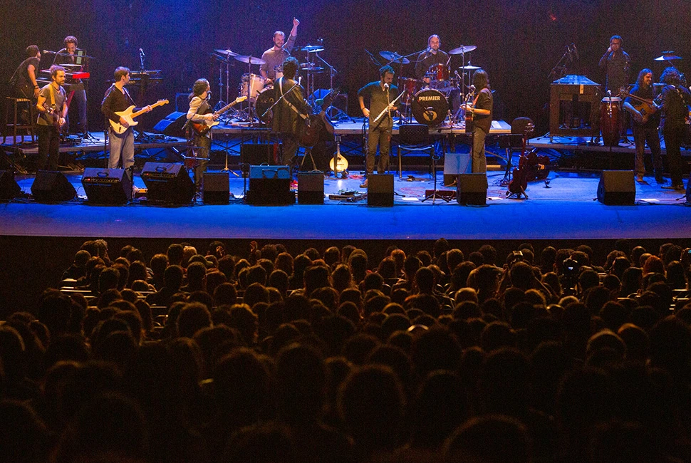 Silhuetas da plateia em primeiro plano assistindo ao show da Orquestra Mineira de Rock no palco iluminado do Sesc Palladium.