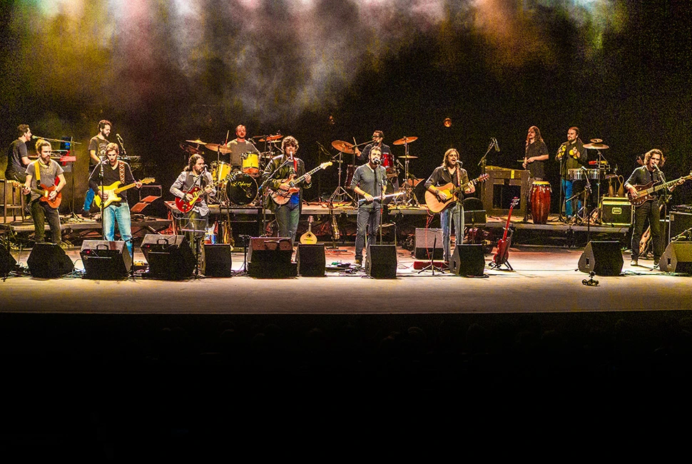 Músicos da Orquestra Mineira de Rock alinhados na frente do palco sob iluminação quente e amarela.