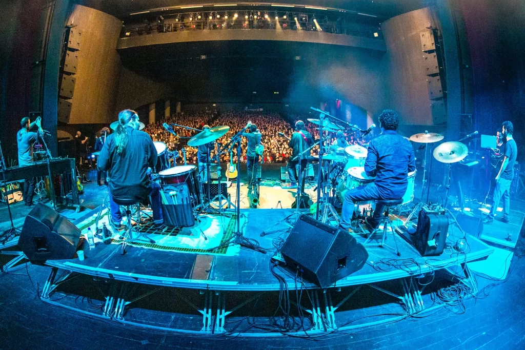 Visão posterior dos músicos da Orquestra Mineira de Rock e da platéia com foco na iluminação azul e profundidade do palco.