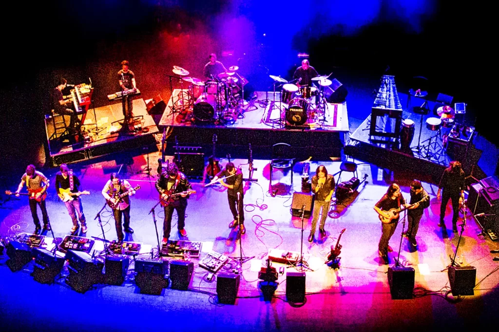 Perspectiva elevada do palco com músicos da Orquestra Mineira de Rock e sombras projetadas sob luz azul.