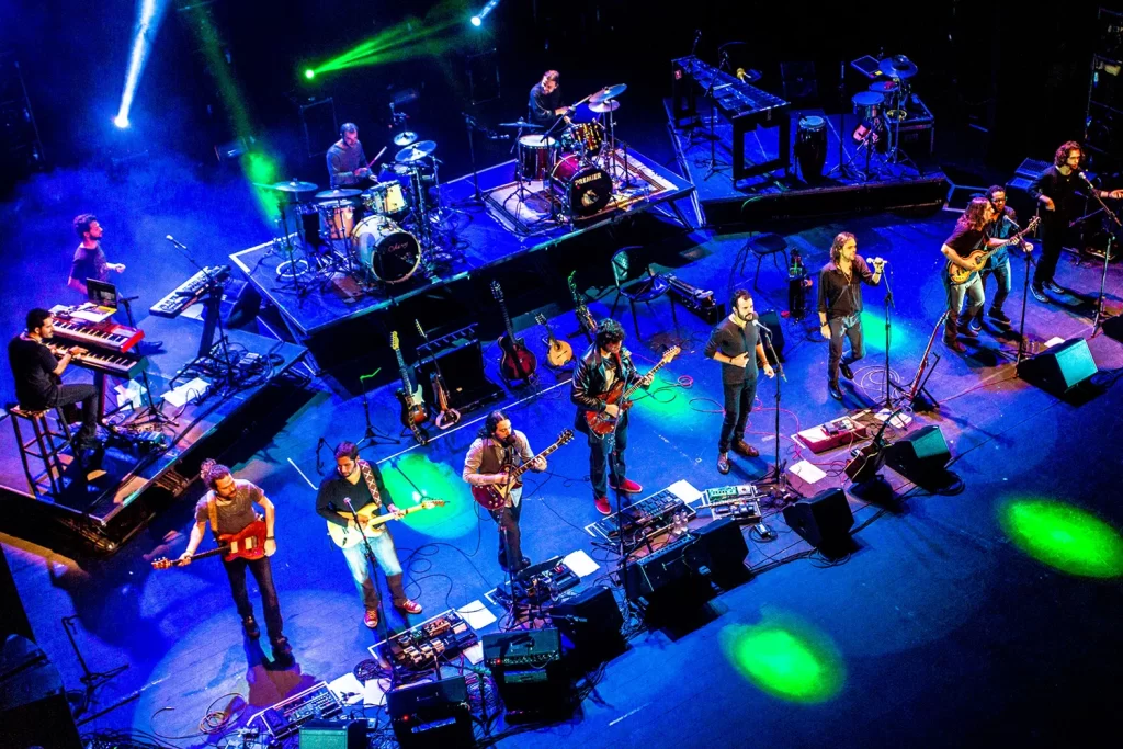 Vista panorâmica superior do palco do Sesc Palladium com os integrantes da Orquestra Mineira de Rock tocando sob iluminação dramática azul e amarela.