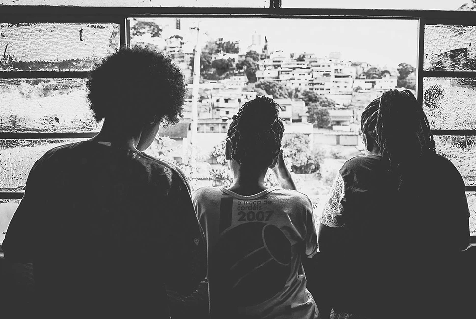 Três pessoas observam a paisagem urbana de Belo Horizonte através de uma grande janela, em silhueta contra a luz, fotografia de Ricardo Laf.