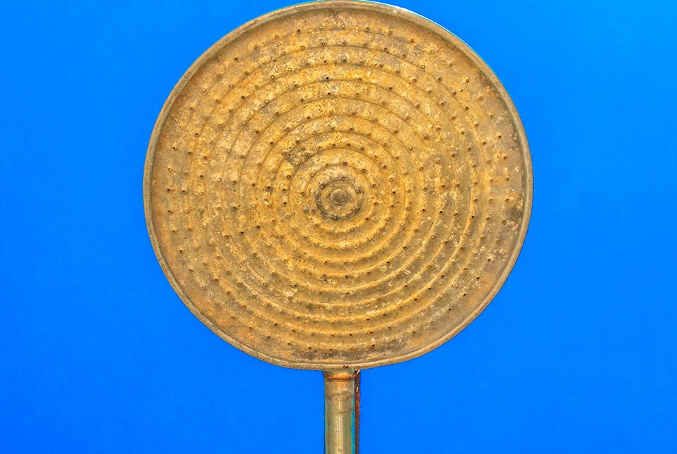 Fotografia em contra-luz de um chuveiro antigo de metal com marcas de ferrugem alaranjada, visto de baixo para cima contra um céu de azul intenso e límpido.