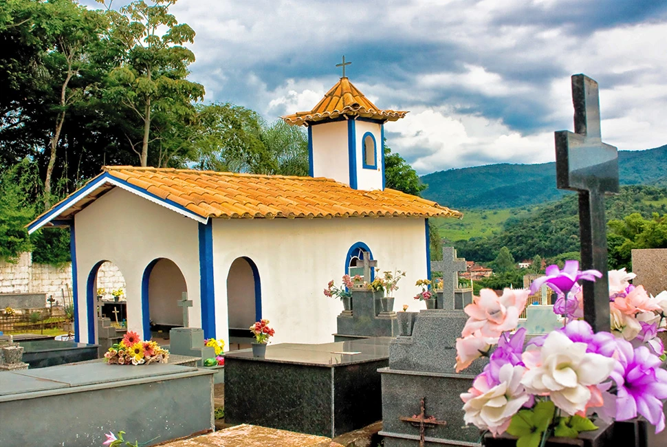 Fotografia de Ricardo Laf mostrando uma pequena capela branca e azul em um cemitério na cidade de Capelinha, Minas Gerais, com flores coloridas em primeiro plano.