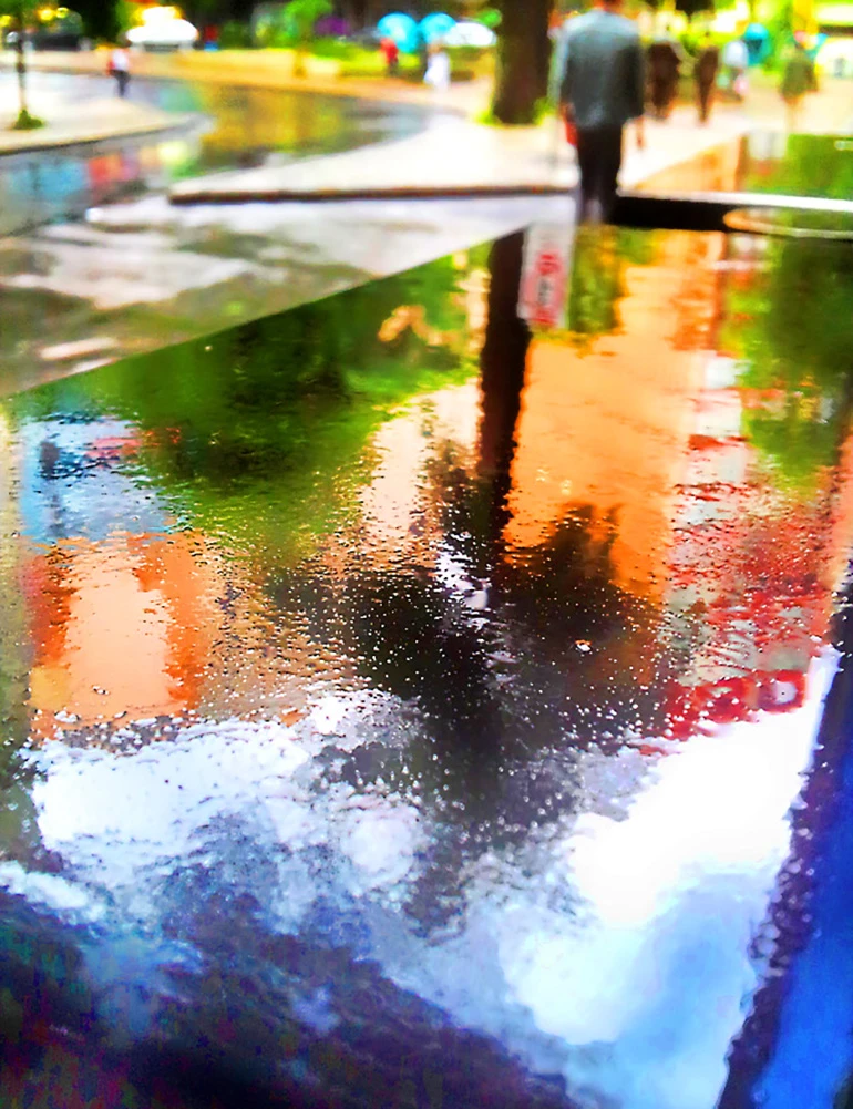 Fotografia de Ricardo Laf capturando o reflexo da arquitetura e de pessoas caminhando na água acumulada sobre uma lixeira metálica no Centro de BH.