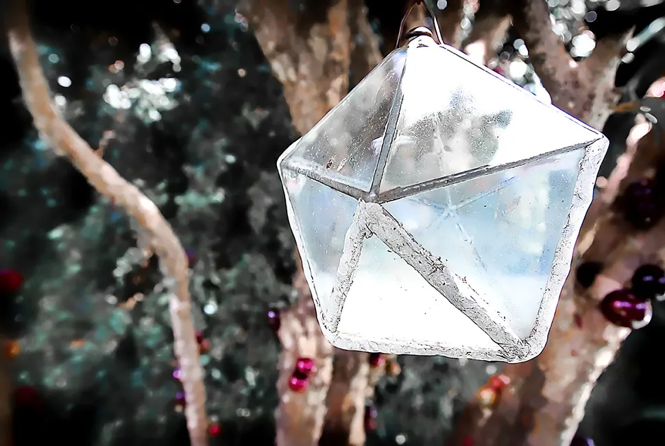 Foto de Ricardo Laf mostrando um enfeite transparente em formato geométrico facetado, pendurado em galhos de uma árvore com iluminação contrastante ao fundo.