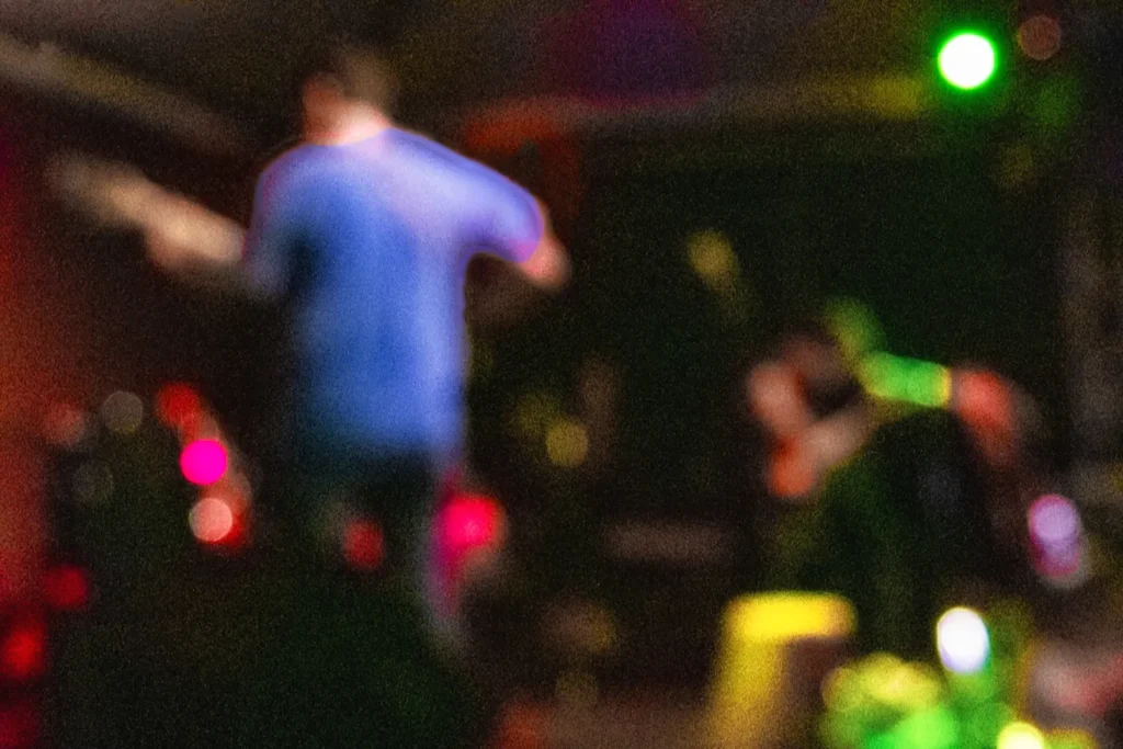 Fotografia autoral propositalmente desfocada e com ruído digital, exibindo um músico em tons de azul e luzes coloridas de palco (bokeh), representando a sinestesia entre som e imagem.