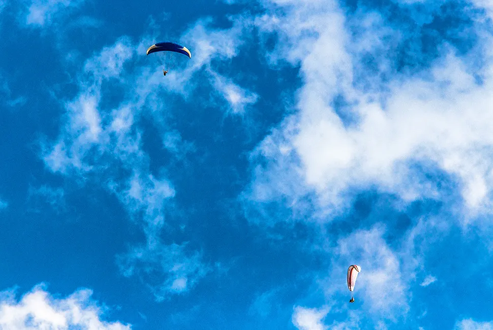 Fotografia de Ricardo Laf mostrando dois paragliders voando sob um céu azul vibrante com nuvens brancas esparsas na Serra da Moeda, Minas Gerais.