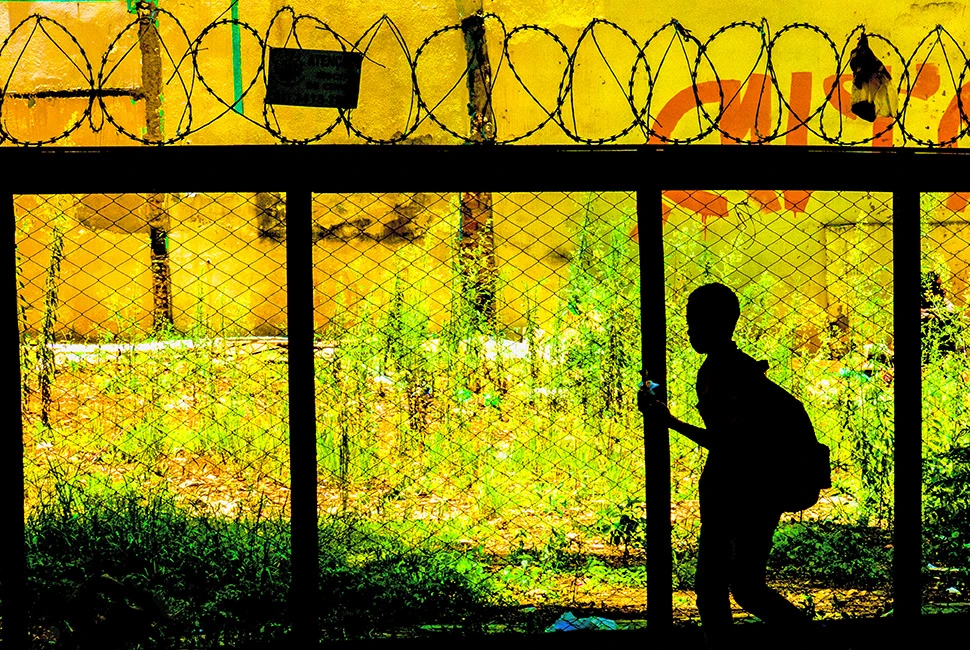 Silhueta de um jovem estudante com mochila, vista através da janela amarela de um ônibus em movimento no centro de Belo Horizonte, com detalhes de grades e reflexos urbanos.