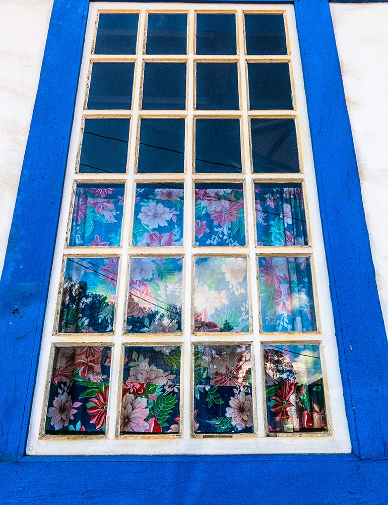 Foto de Ricardo Laf focada no detalhe de uma janela colonial azul em Lavras Novas, Minas Gerais, com vidros pequenos e cortinas aparentes.