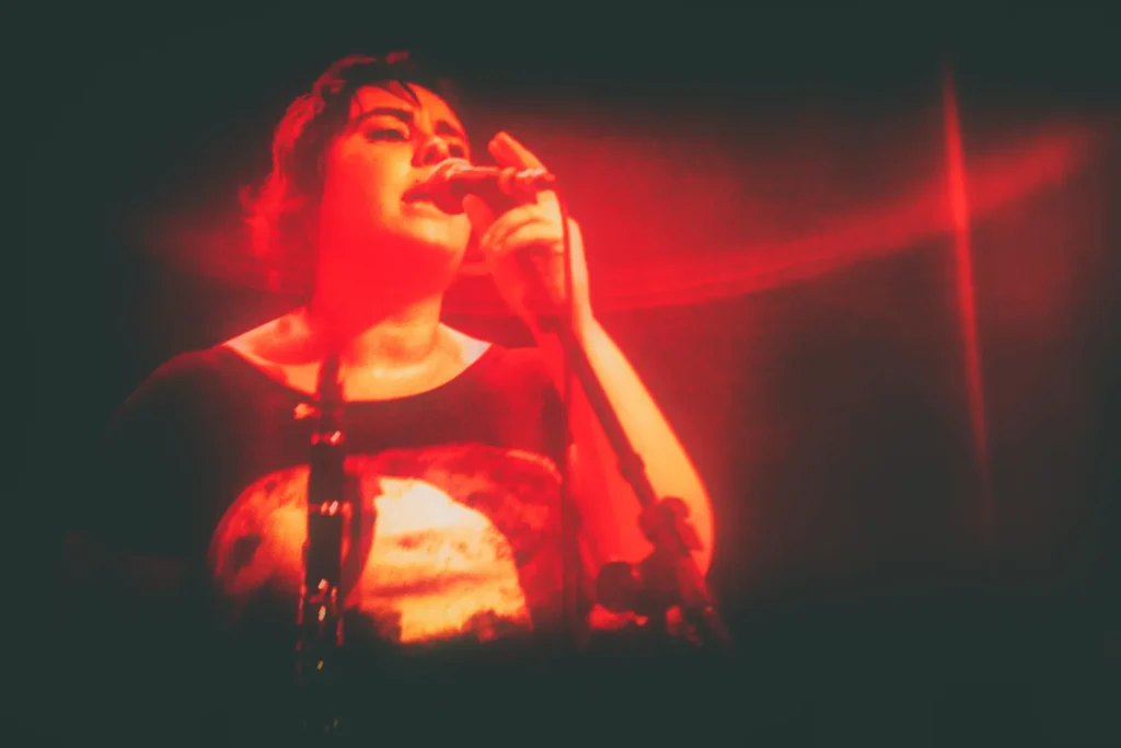 Fotografia de Ricardo Laf capturando a expressividade intensa de Juliana Perdigão sob luzes vermelhas dramáticas, destacando a energia e a emoção do momento musical.