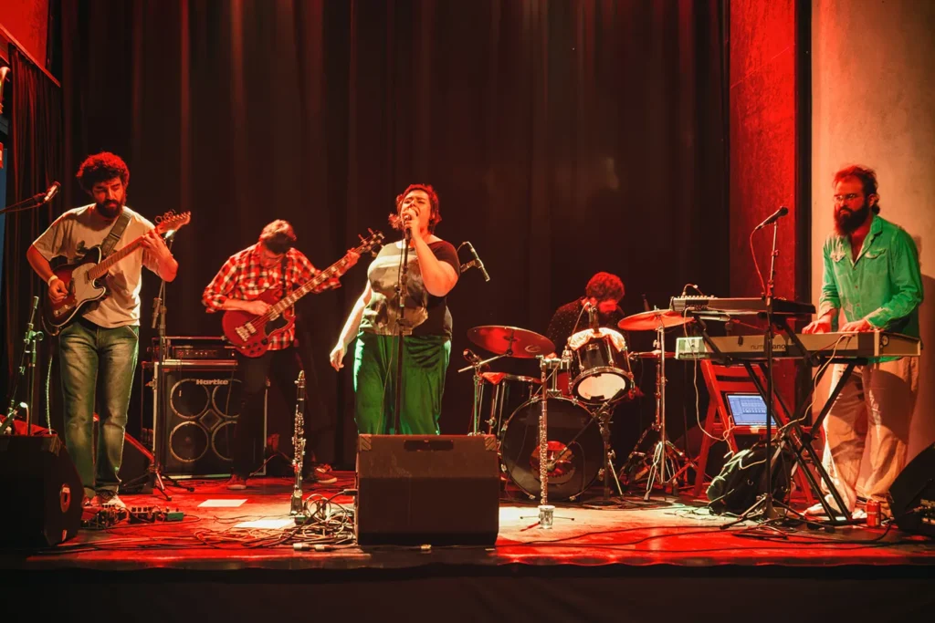 Fotografia de Ricardo Laf registrando Juliana Perdigão e sua banda em performance no palco, destacando a composição coletiva sob luzes dramáticas e atmosfera artística.