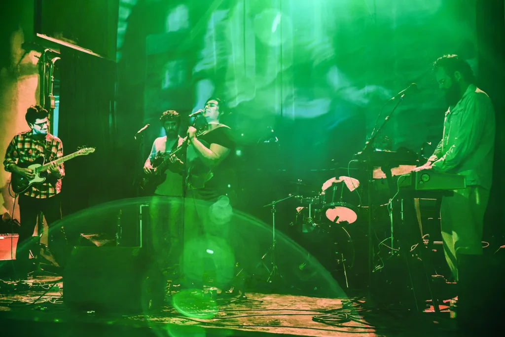 Fotografia de Ricardo Laf capturando Juliana Perdigão e banda num momento de grande energia cénica, com luzes verdes vibrantes que criam uma atmosfera experimental e imersiva.