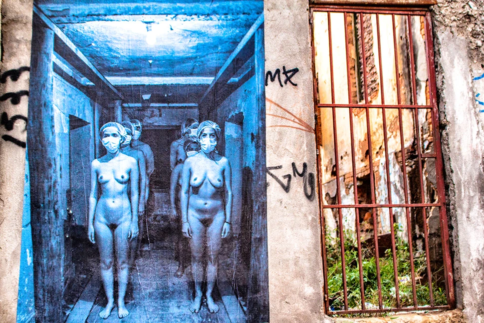 Fotografia colorida de uma parede externa no centro de Belo Horizonte com intervenções de arte urbana (graffiti) ao lado de uma janela com grade enferrujada e textura de parede descascada.