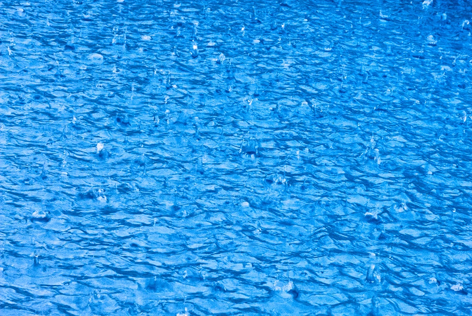 Fotografia em plano fechado da superfície de uma piscina com água azul vibrante, exibindo texturas de pequenas ondas e padrões de refração da luz solar.