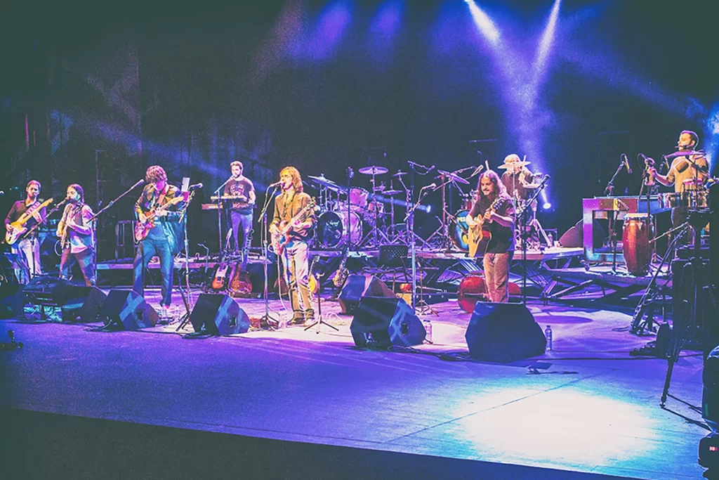 Integrantes da Orquestra Mineira de Rock tocando guitarras e instrumentos de percussão no palco do Sesc Palladium sob luzes azuis dramáticas.