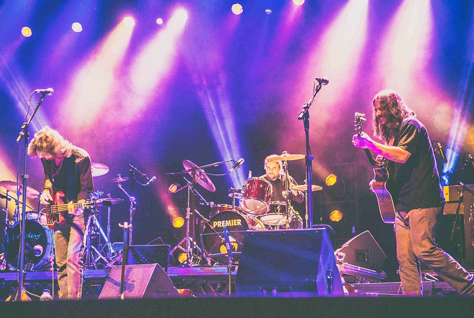 Close médio de um músico da Orquestra Mineira de Rock tocando guitarra em um momento de solo, banhado por luzes brancas e azuis.