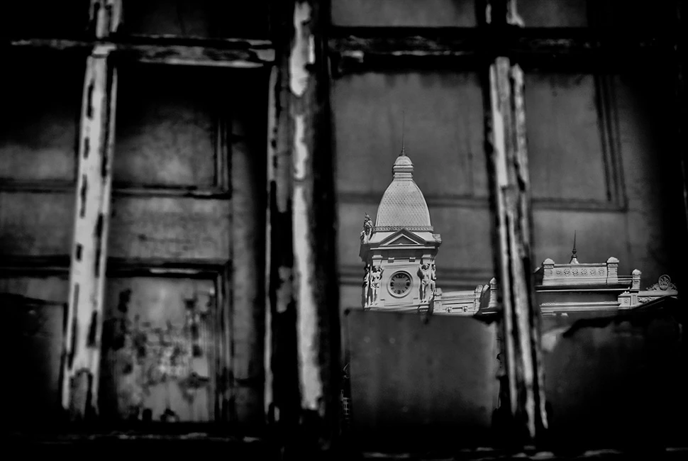 Fotografia em preto e branco de uma janela antiga de trem ou edificação na Praça da Estação, em Belo Horizonte, com reflexos que misturam a estrutura metálica e o ambiente urbano.