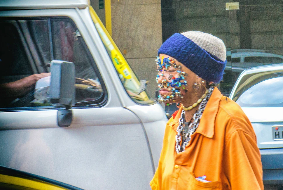 Retrato de perfil de uma mulher idosa usando uma touca azul com detalhes coloridos, diversos piercings no rosto e uma camisa laranja vibrante, em uma rua movimentada do centro de Belo Horizonte.
