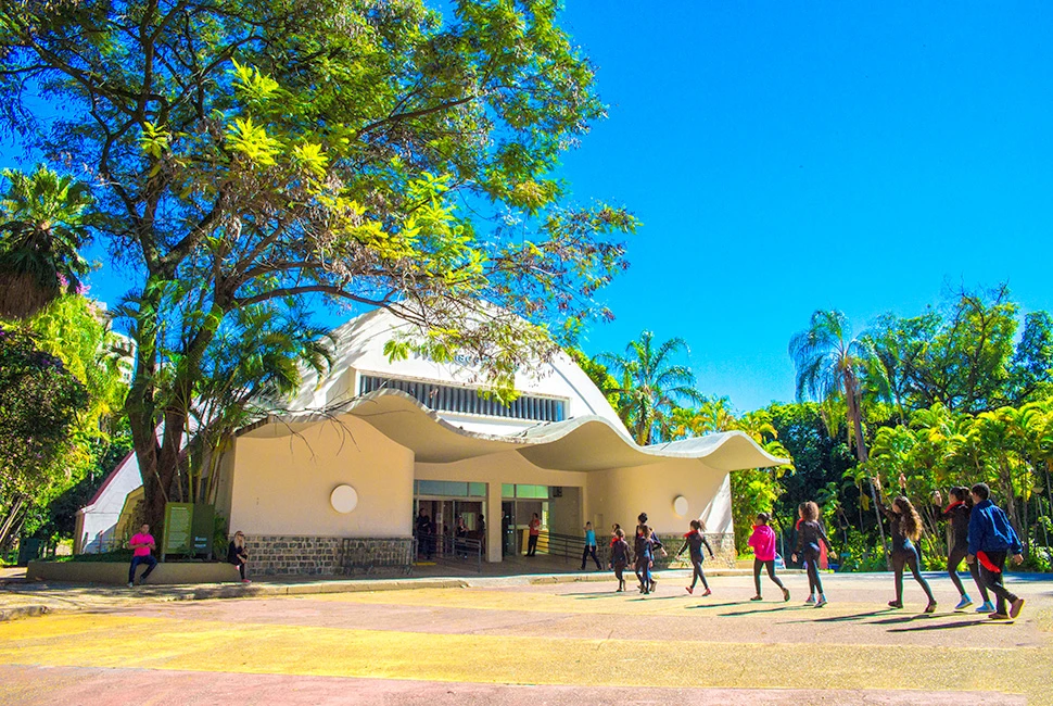 Fachada branca e moderna do Teatro Francisco Nunes em Belo Horizonte, cercada pela vegetação densa e árvores do Parque Municipal sob um céu azul claro.