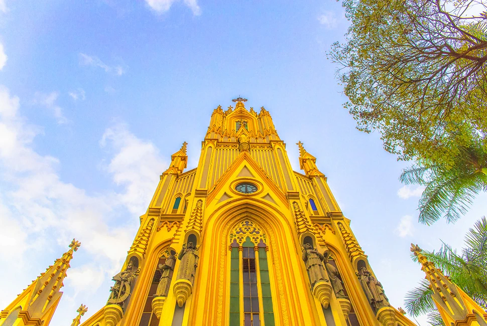 Fotografia em ângulo baixo da Basílica de Nossa Senhora de Lourdes em Belo Horizonte, destacando sua fachada amarela de estilo neogótico contra um céu azul e árvores laterais.