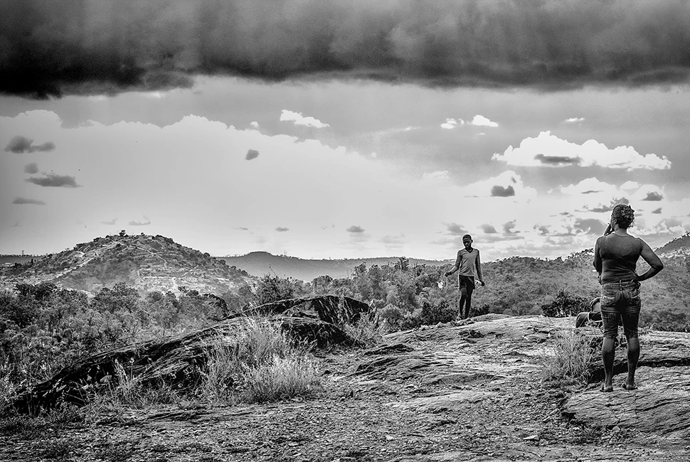 Fotografia em preto e branco de uma pedreira em Santa Luzia, Minas Gerais, mostrando figuras humanas distantes em um terreno rochoso sob um céu com nuvens densas e carregadas.