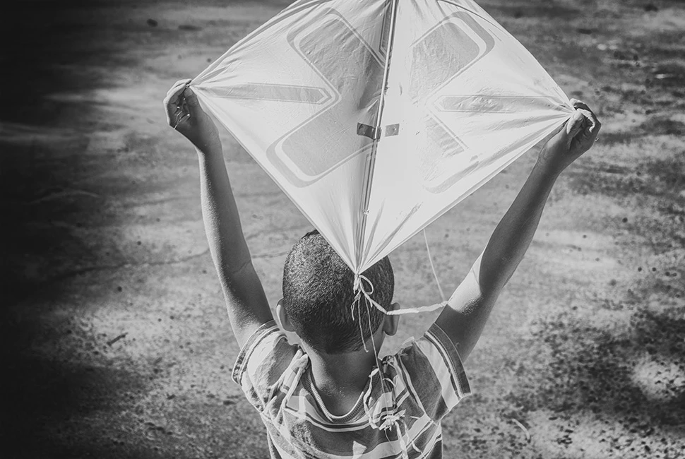 Fotografia em preto e branco de costas de um menino segurando uma pipa (papagaio) contra um céu nublado em Santa Luzia, Minas Gerais.