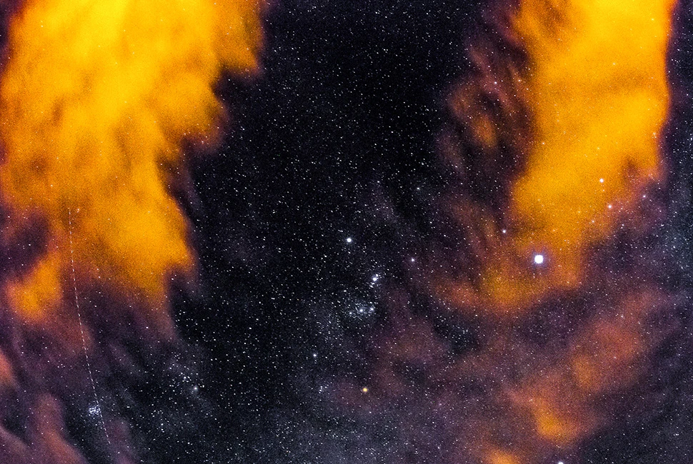 Foto de Ricardo Laf capturada em longa exposição, mostrando o céu estrelado e nuvens iluminadas em tons de dourado e violeta em Lagoa Santa, Minas Gerais.