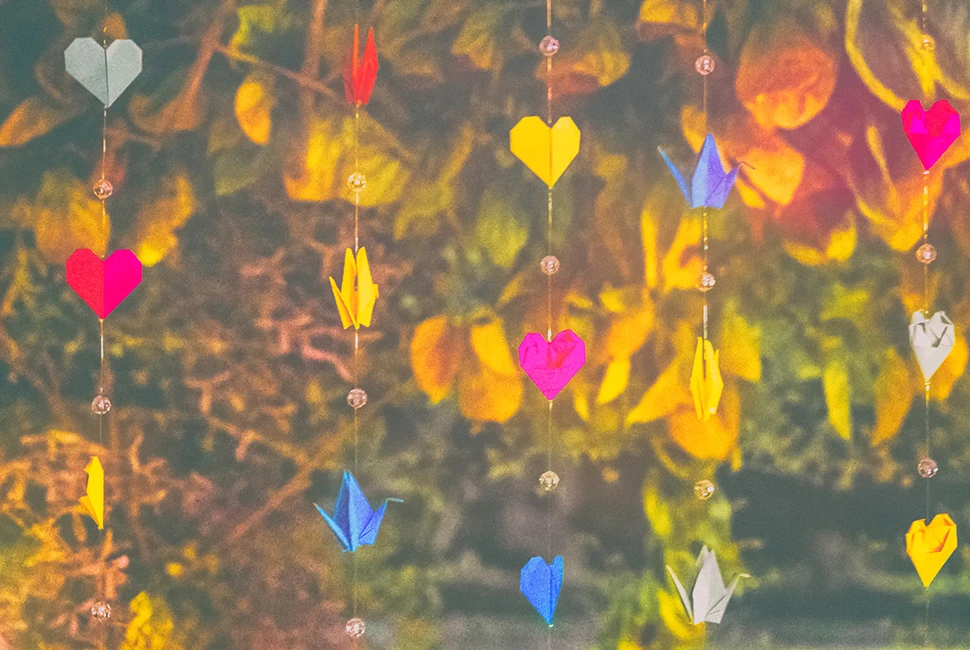 Fotografia de detalhes de enfeites em formato de coração, coloridos e pendurados, com um fundo desfocado de vegetação e luz natural.
