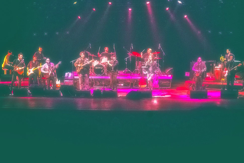 Fotografia de palco da Orquestra Mineira de Rock com iluminação intensa em tons de rosa e magenta. Músicos em performance coletiva no Teatro Bradesco.