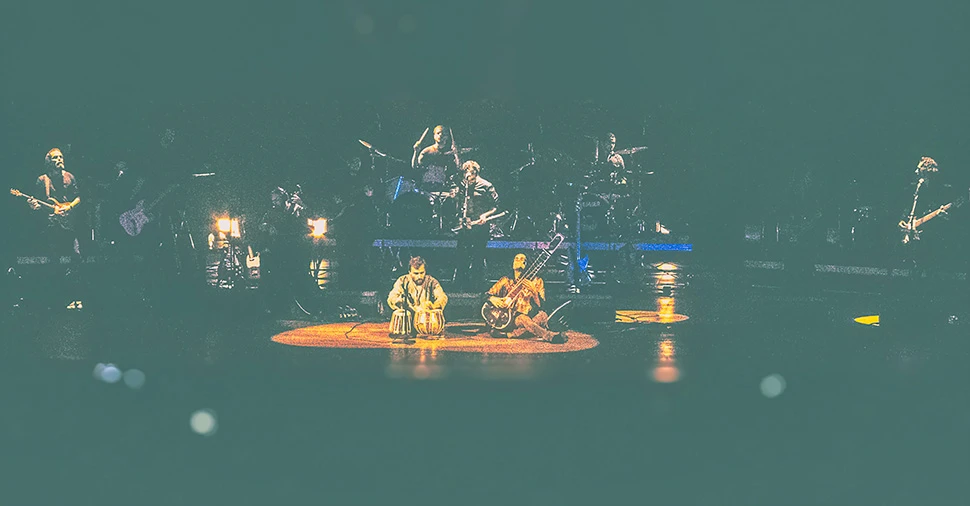 Fotografia de plano geral intimista da Orquestra Mineira de Rock. Músicos no palco do Teatro Bradesco sob iluminação suave e tons quentes.