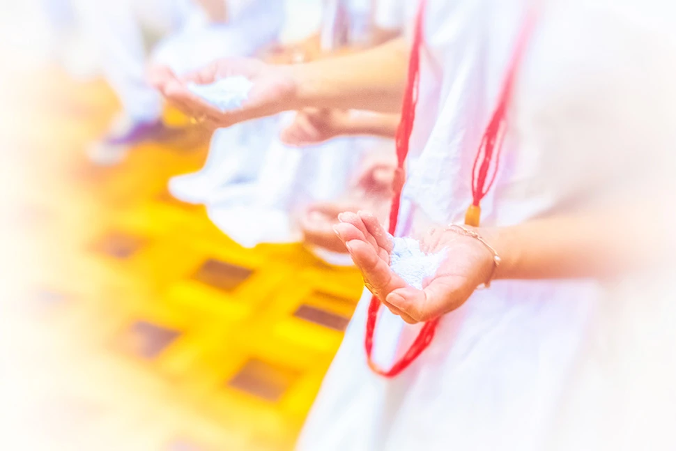 Fotografia com foco suave e iluminação clara, mostrando mãos em concha segurando um pequeno objeto simbólico ou sagrado, com vestes brancas e detalhes em vermelho ao fundo.