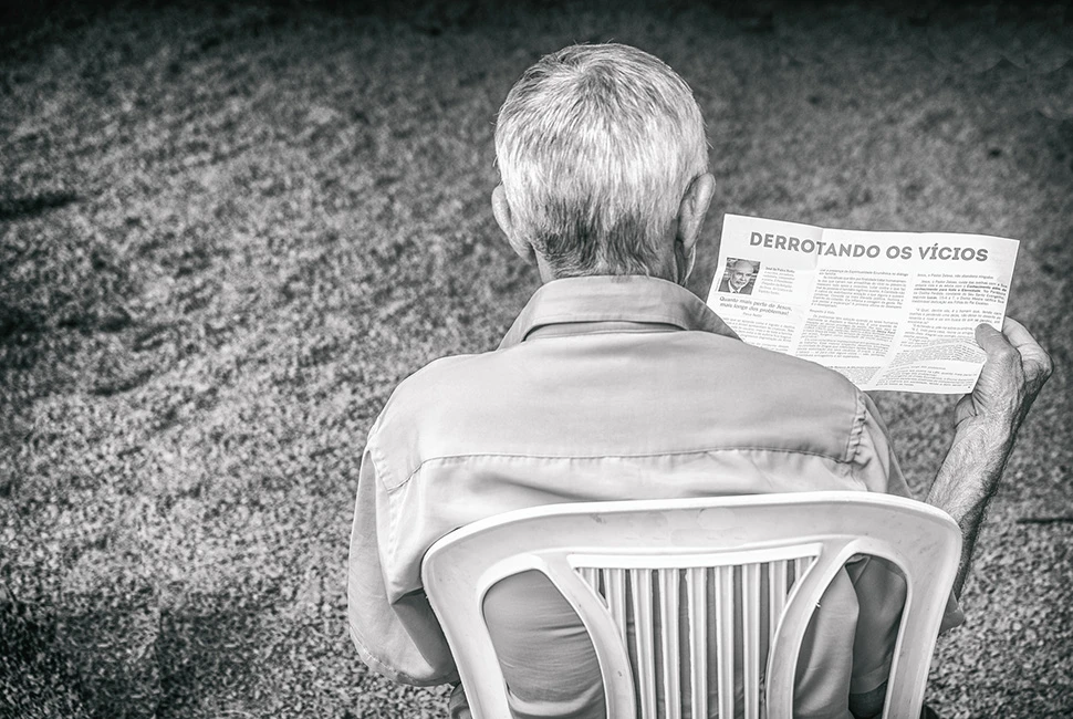 Fotografia em preto e branco mostrando um homem sentado de costas, lendo um jornal ou panfleto com a manchete "Derrotando os Vícios", em um ambiente externo em Belo Horizonte.