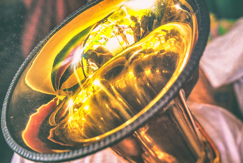 Fotografia em close focando na campana de um trombone de metal dourado, destacando os reflexos de luz na superfície curva e as texturas do bocal e das chaves.
