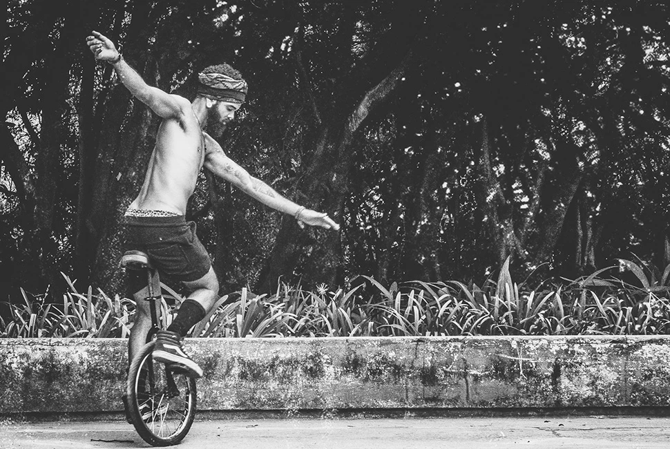 Fotografia em preto e branco de um homem sem camisa equilibrando-se em um monociclo sobre uma mureta de pedra, com vegetação densa ao fundo, em Belo Horizonte.